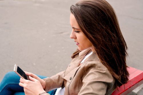 Free A woman in a brown jacket using a smartphone while sitting on a bench outdoors. Stock Photo