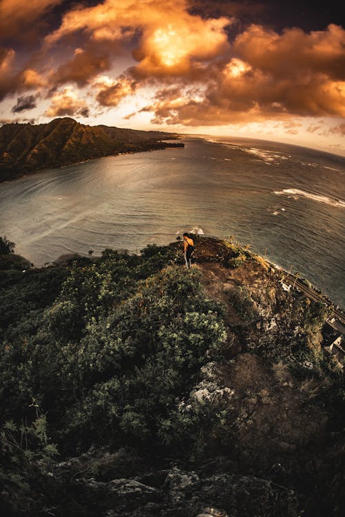 匿名的徒步旅行者在日落期间在风景如画的海中达到岩石峭壁的顶峰