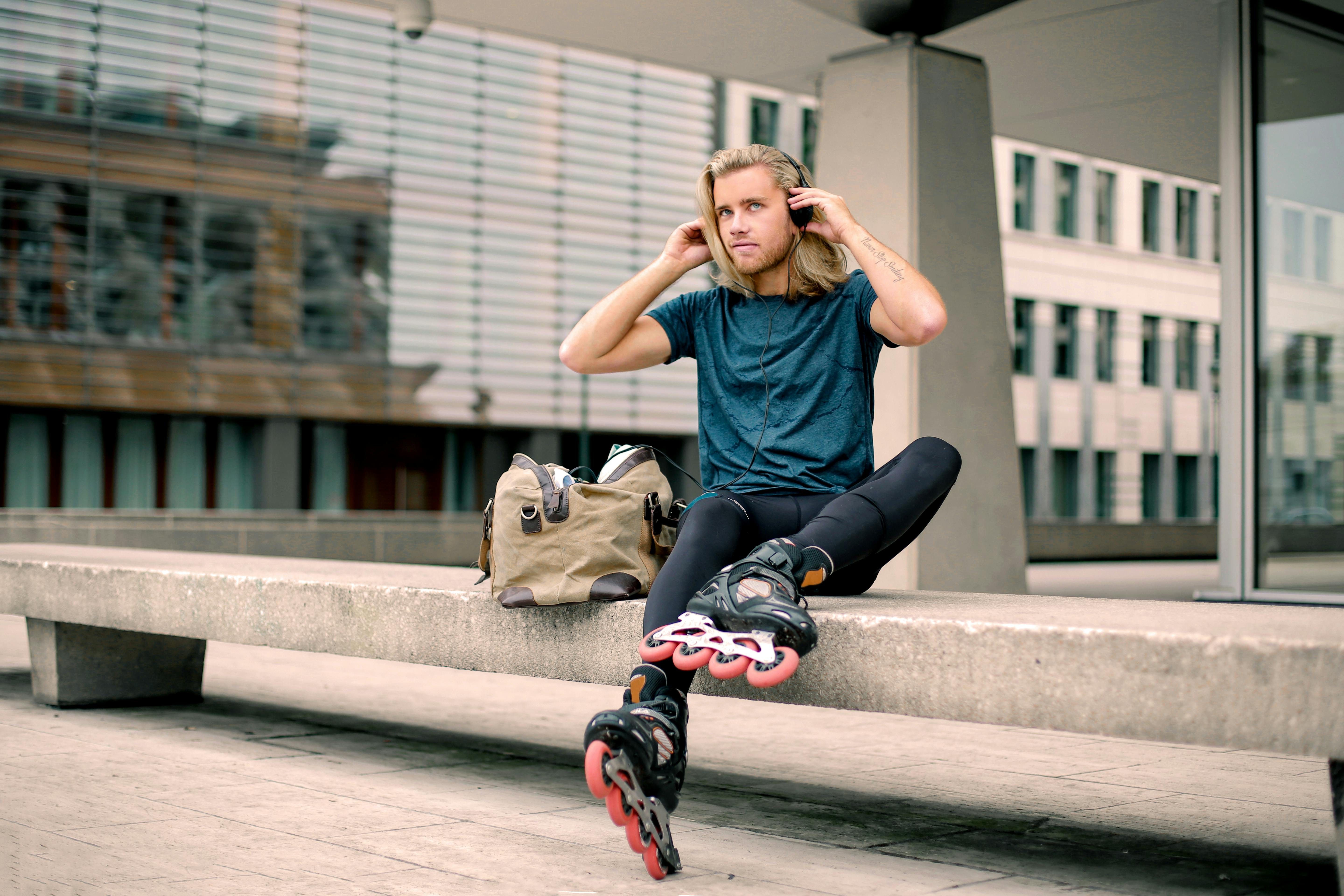 Adult man wearing headphones and rollerblades, sitting on a bench in a modern urban environment.