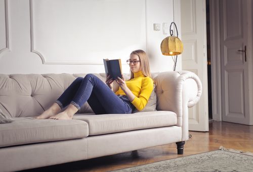 Free Young woman enjoying a leisure time reading a book on a cozy beige sofa in a bright living room. Stock Photo