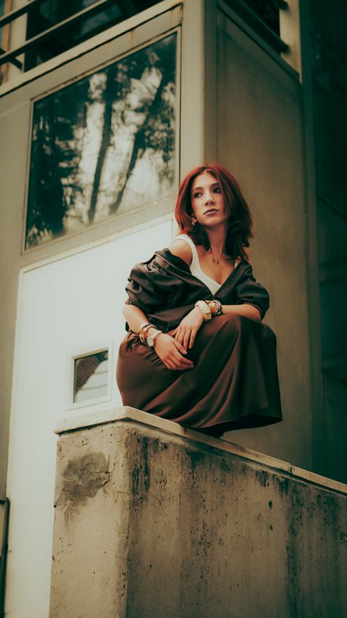 Free Fashionable young woman in elegant dress posing confidently on an urban balcony with a modern building backdrop. Stock Photo