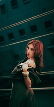 Elegant woman with red hair poses against a city building backdrop, exuding modern fashion style.
