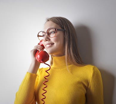 Woman in yellow sweater smiling while talking on a vintage red phone against a white wall.