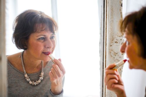 Free Elegant senior woman applying lipstick while looking in the mirror indoors. Stock Photo