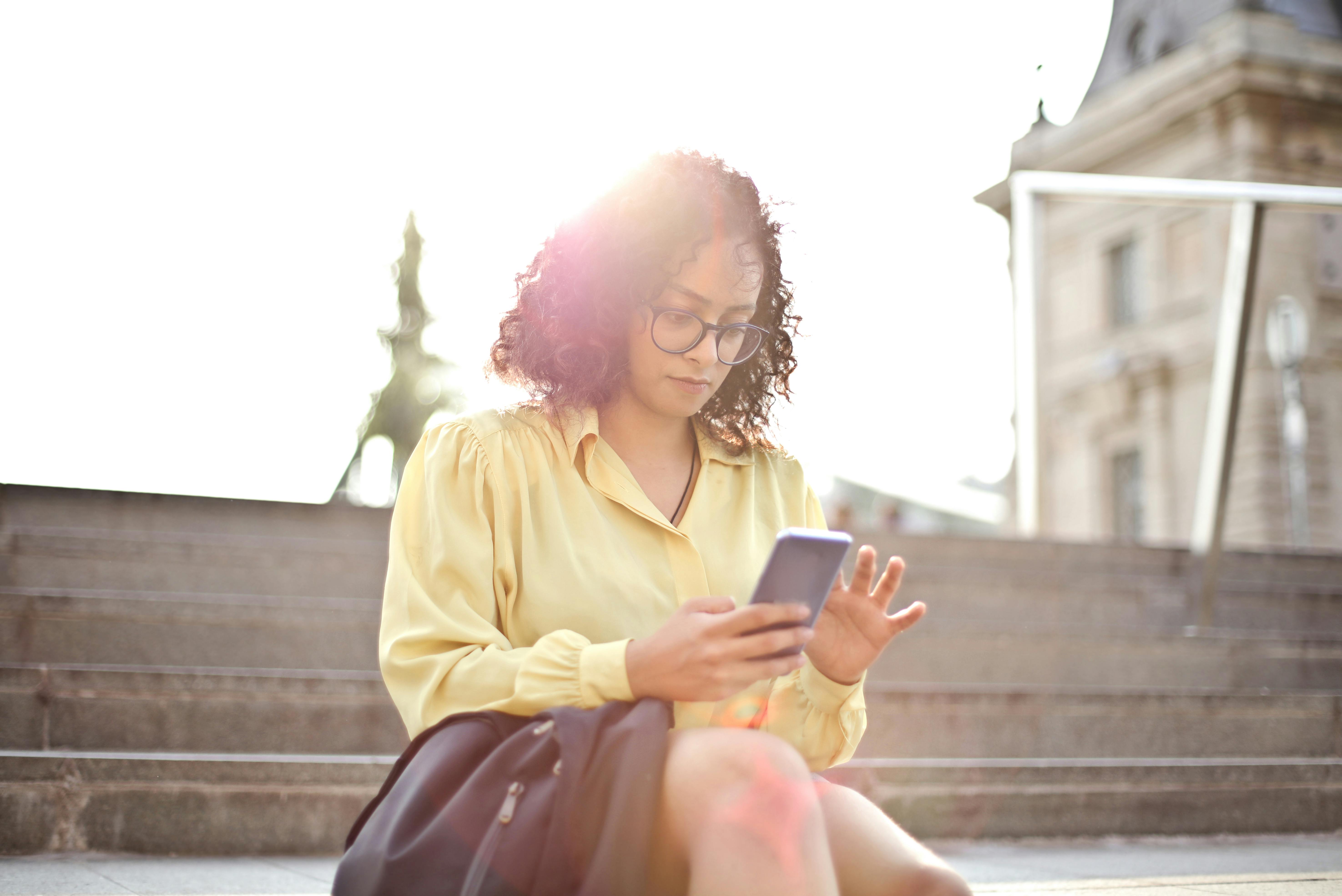 selective focus photo of woman in yellow dress shirt sitting on stairs using her phone