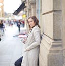 热闹的城市街道上，一位穿着风衣、面带微笑的女人正靠在墙上使用智能手机