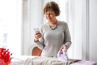 Happy middle aged woman in casual clothes focusing on screen and interacting with smartphone while ironing clothes in light living room of modern apartment