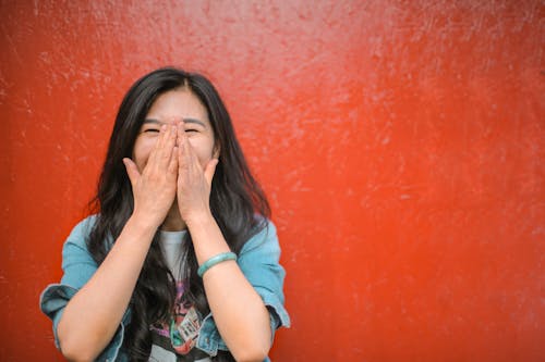 Young overjoyed Asian female in casual outfit covering face with hands and laughing while standing against vibrant orange wall