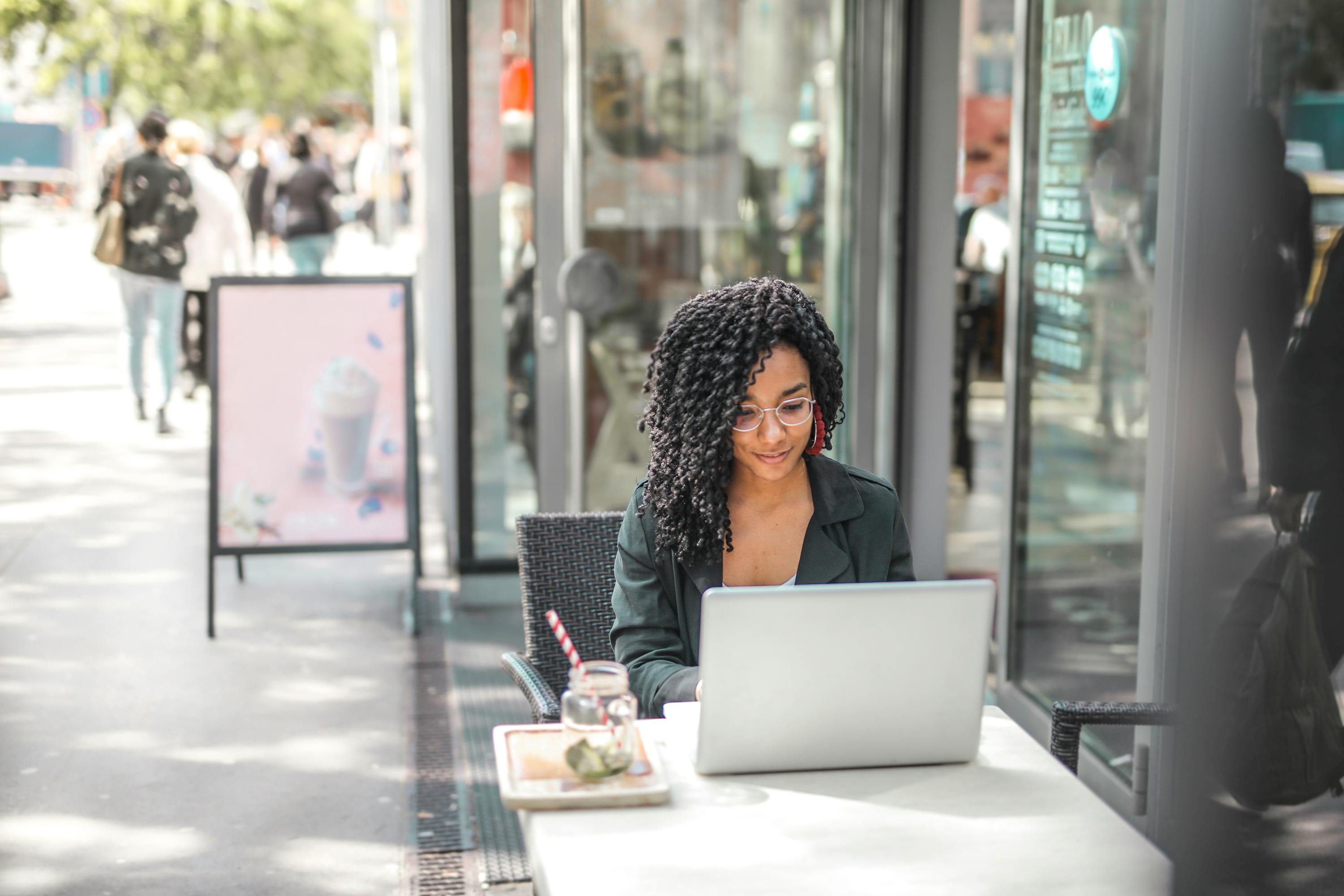 ethnic young woman using laptop while having tasty beverage in modern street cafe