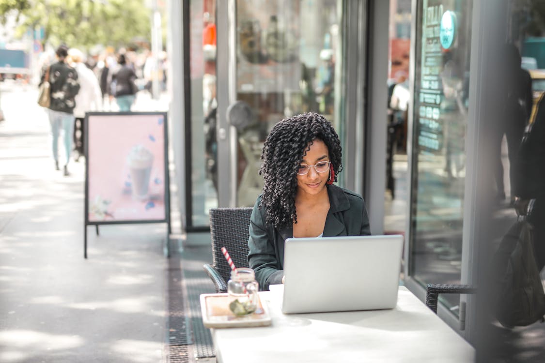 Free High angle of pensive African American female freelancer in glasses and casual clothes focusing on screen and interacting with netbook while sitting at table with glass of yummy drink on cafe terrace in sunny day Stock Photo