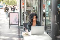 High angle of pensive African American female freelancer in glasses and casual clothes focusing on screen and interacting with netbook while sitting at table with glass of yummy drink on cafe terra...