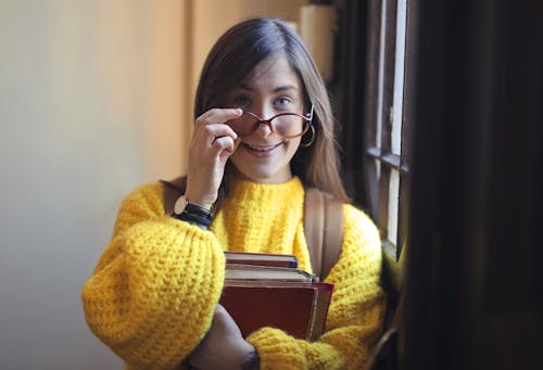 Portrait Photo of Smiling Woman in Yellow Knit Sweater and Black Framed Glasses Standing by Window While Carrying Books