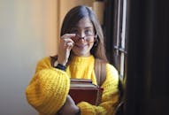 一个开朗的女人戴着眼镜，拿着书，站在室内的窗户旁