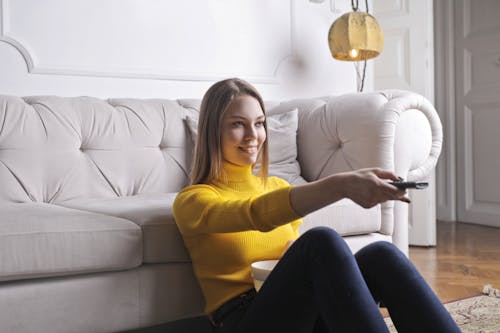 Joyful millennial female in casual clothes with bowl of snack using remote controller while sitting on floor leaning on sofa and watching movie in cozy light living room with luxury interior