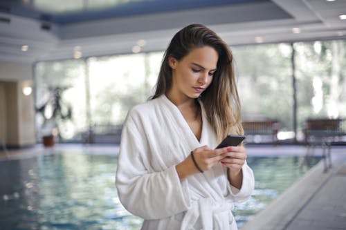 Selective Focus Photo of Woman in White Robe Standing Poolside While Using Her Smartphone