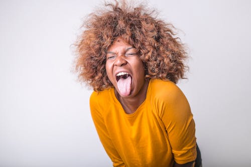 Free Red haired African American female in vivid yellow sweater standing with eyes closed and tongue out against gray background in modern studio Stock Photo