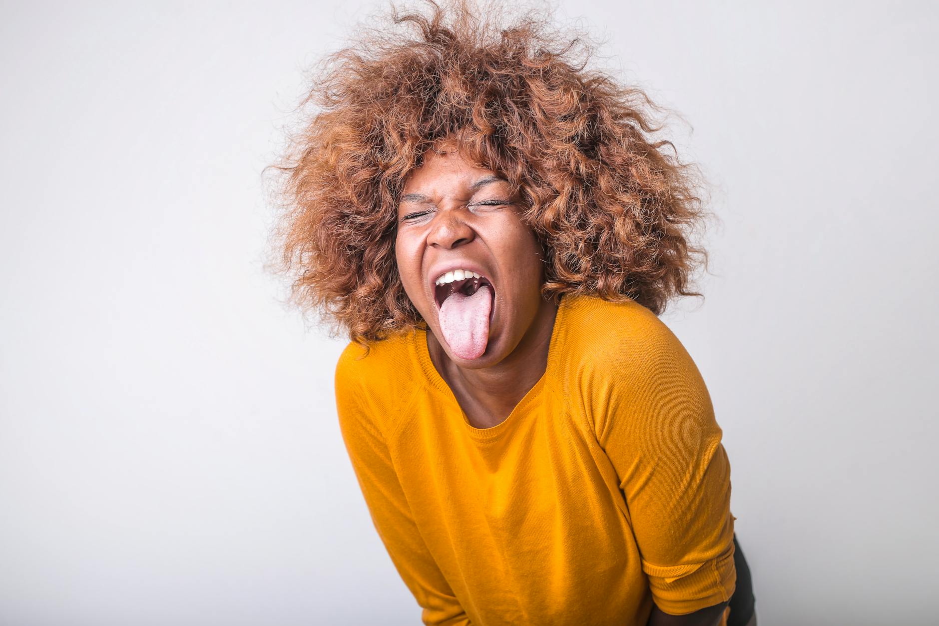 Red haired African American female in vivid yellow sweater standing with eyes closed and tongue out against gray background in modern studio
