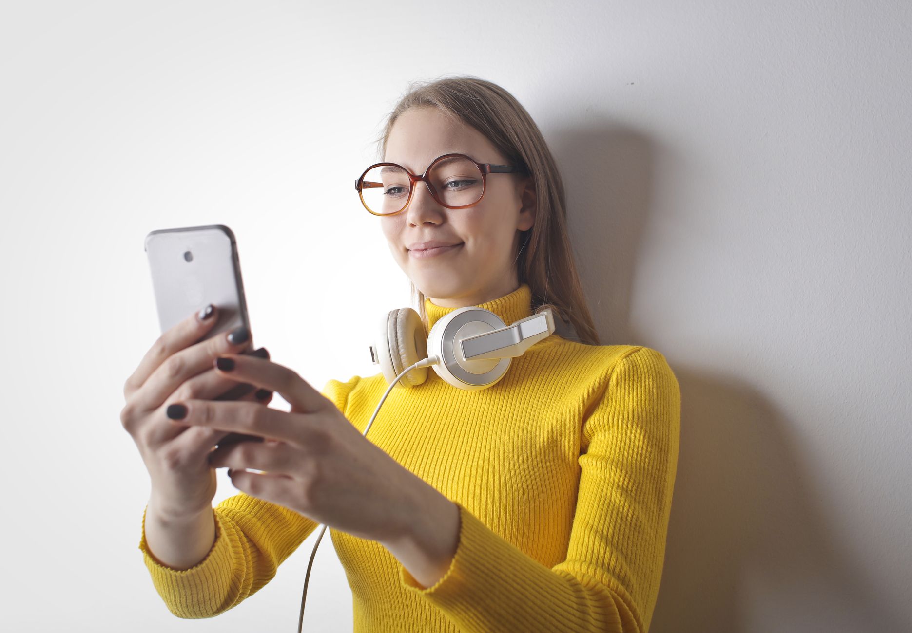 Young woman in yellow sweater taking a selfie with a smartphone, wearing eyeglasses and headphones.