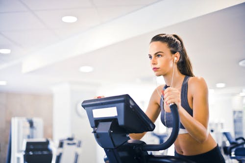 Serious fit woman in earphones and activewear listening to music and running on treadmill in light contemporary sports center