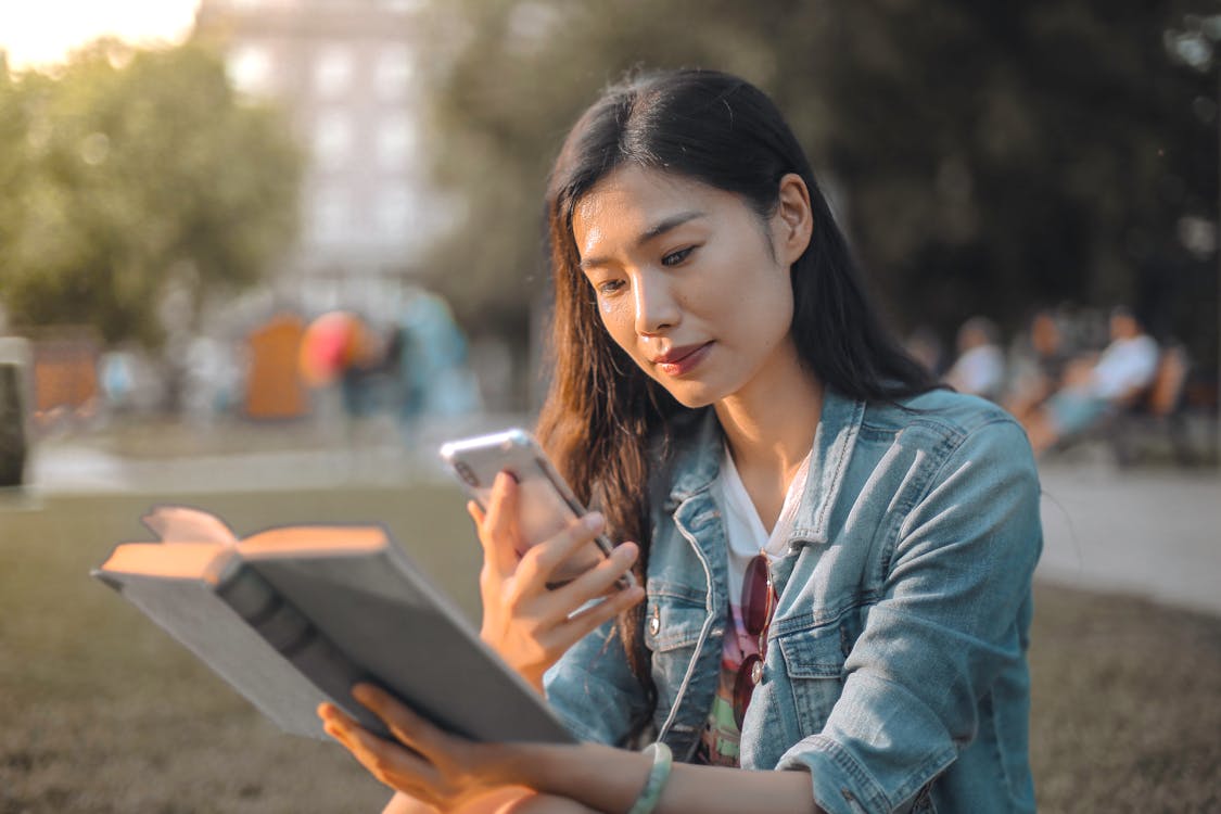 Free Selective Focus Photo of Woman in Blue Denim Jacket Using Her Phone While Holding a Book Stock Photo