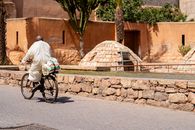 A man rides a bicycle along a stone path near a traditional Moroccan wall under bright sunlight.