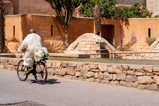 A man rides a bicycle along a stone path near a traditional Moroccan wall under bright sunlight.