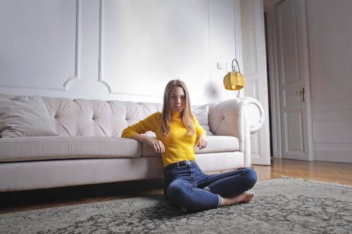 白いソファに寄りかかって足を組んで床に座っている黄色いタートルネックのセーターと青いデニムジーンズの女性の写真