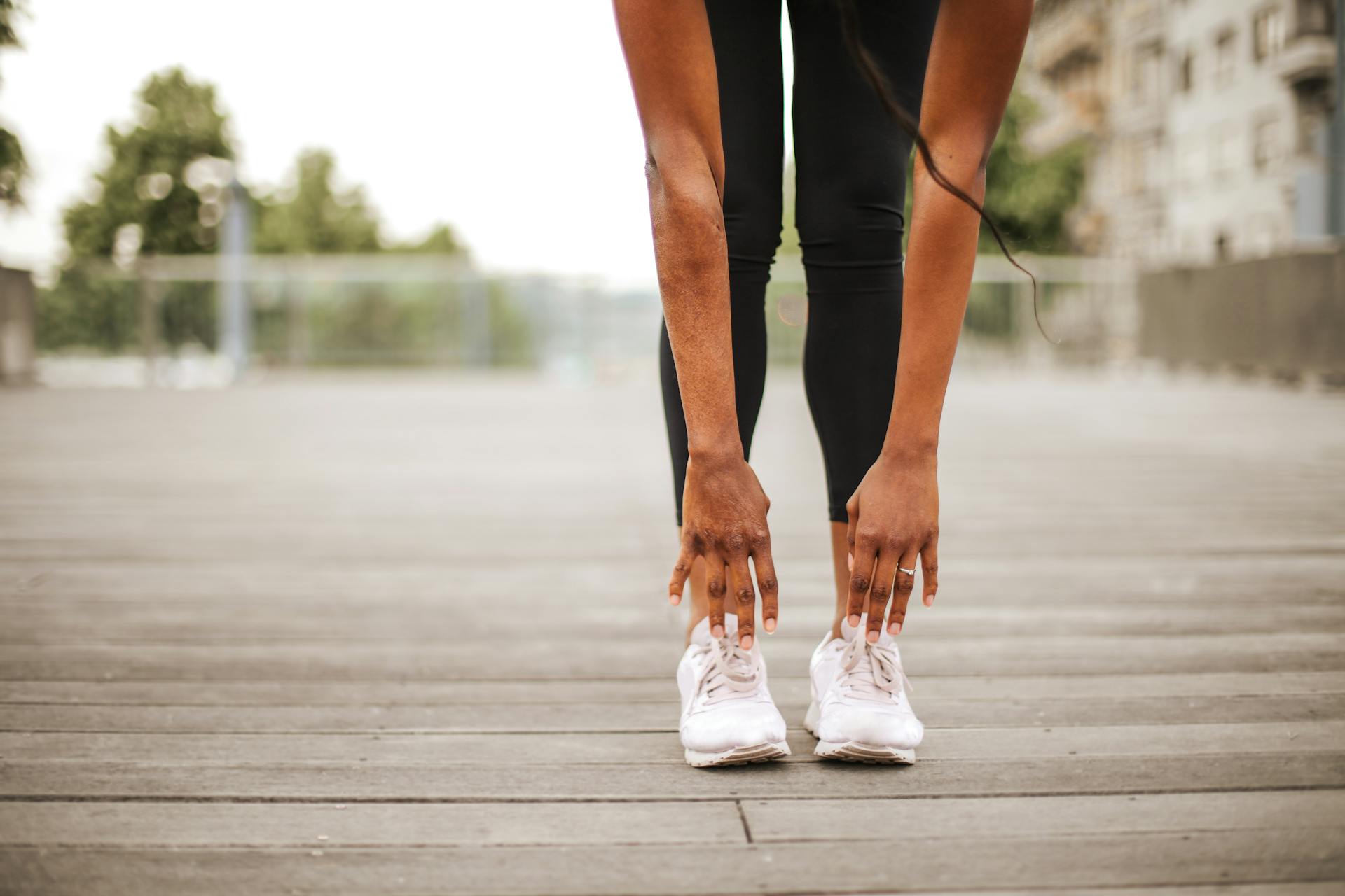 From below crop slender female athlete in sportswear and white sneakers doing standing forward bend exercise for stretching body on wooden floor of street sports ground against blurred urban environment in daytime