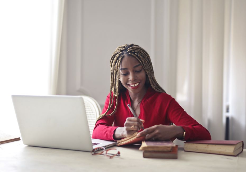A woman with braided hair wearing a red top working at a desk with a laptop and books. A woman with braided hair wearing a red top working at a desk with a laptop and books.