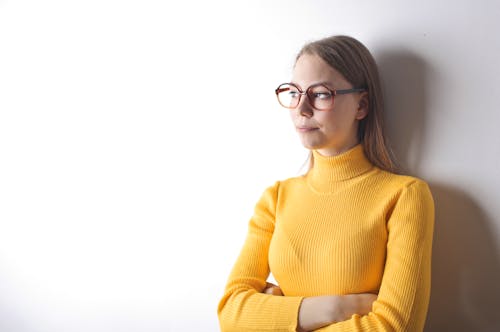 Free Portrait Photo of Woman in Yellow Turtleneck Sweater and Black Framed Eyeglasses Standing In Front of White Wall While Looking Away Stock Photo