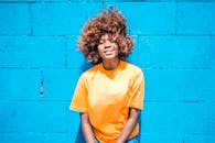 一位留着卷发的开朗女人，穿着休闲的橙色衬衫，靠在蓝色的墙壁上灿烂地微笑着