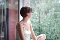 侧视图：清晨，一位穿着休闲睡衣、冷静体贴的年轻女性坐在窗台上，透过窗户望着绿色的森林景色