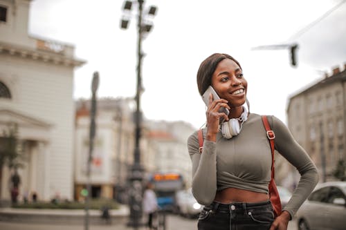 Free Selective Focus Photo of Smiling Woman in Gray Long Sleeve Shirt and Black Denim Jeans Standing While Talking on the Phone Stock Photo