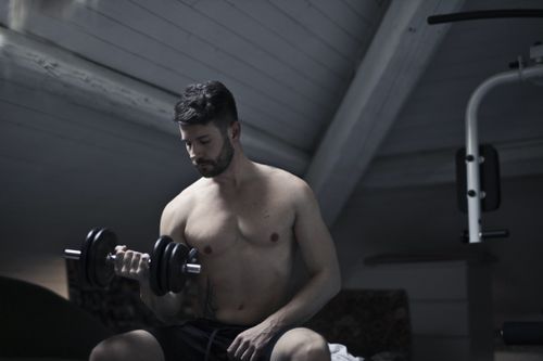 Free From below of muscular shirtless athletic male sitting on bench and doing bicep curls with dumbbell during intense training in dark studio Stock Photo