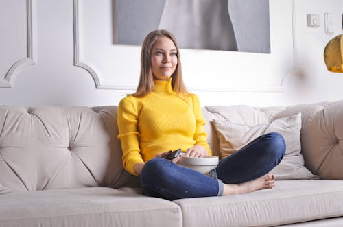 Positive young female in casual clothes sitting on cozy sofa with bowl of snacks and watching interesting film while spending time at home