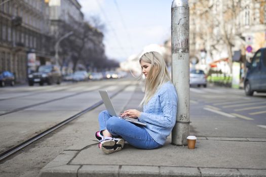 Blonde woman sitting on city street with laptop, embracing urban lifestyle and technology.