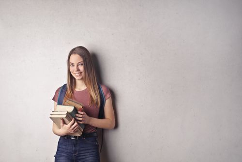 Free A young woman standing against a white wall, smiling while holding books. Casual attire and backpack enhance the academic vibe. Stock Photo