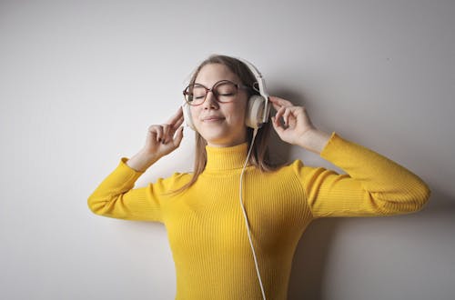 Free Portrait Photo of Woman in Yellow Turtleneck Sweater and Brown Framed Eyeglasses Standing In Front of White Wall With Her Eyes Closed Wearing White Headphones Stock Photo
