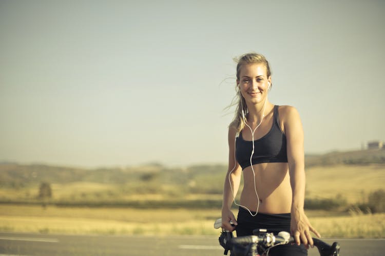 Selective Focus Photo Of Smiling Woman In Black Active Wear With Earphones On  Standing Next To Her Bicycle By The Side Of The Road