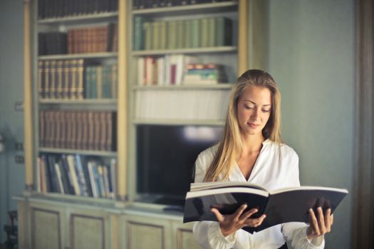 A woman in a white top reading a book in a home library, surrounded by bookshelves.
