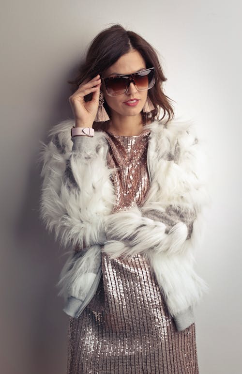 Foto De Mulher Com Casaco De Pele Branca, Vestido Brilhante E óculos De Sol Em Pé Na Frente De Parede Cinza