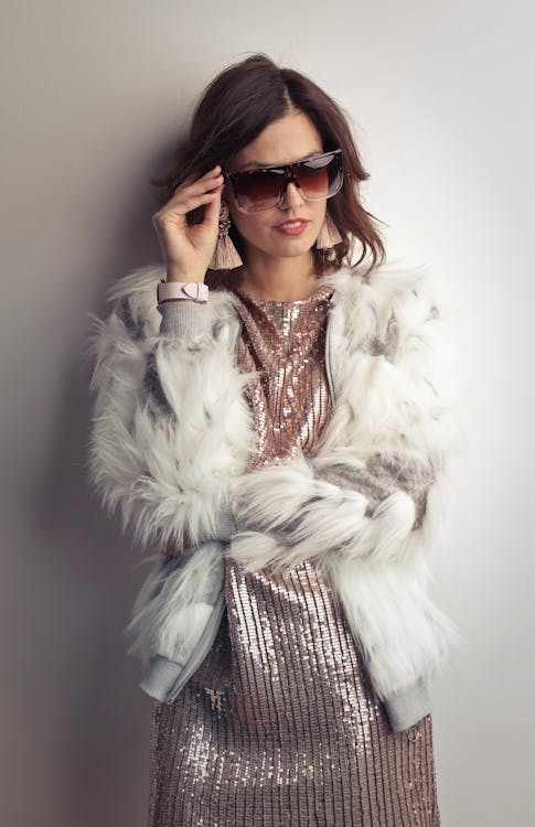 Photo of Woman in White Fur Coat, Shiny Dress, and Sunglasses Standing In Front of Gray Wall