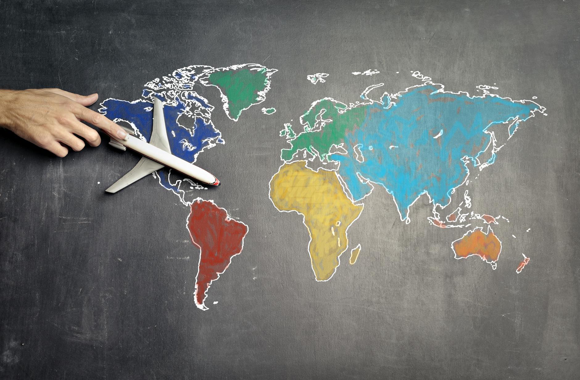 Top view of crop anonymous person holding toy airplane on colorful world map drawn on chalkboard