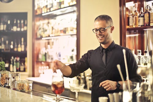 Happy young male barkeeper standing at counter and preparing alcohol cocktail for order while working in modern pub
