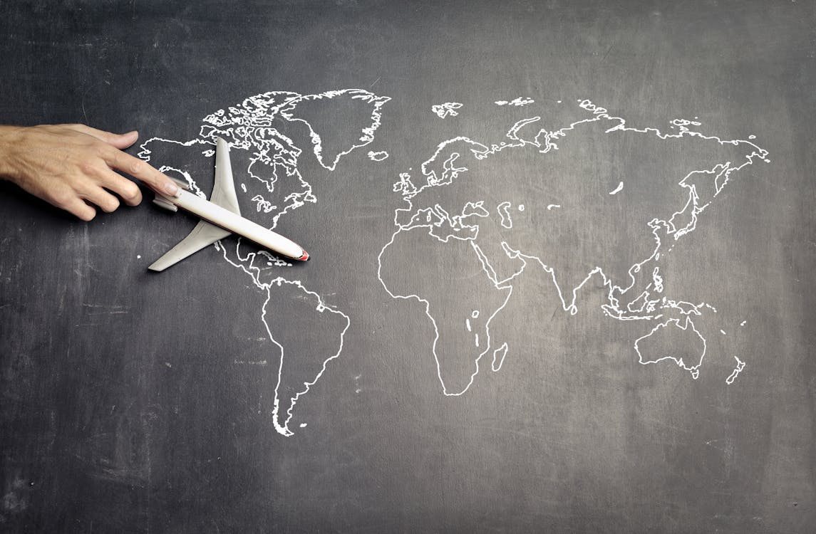 From above of crop anonymous person driving toy airplane on empty world map drawn on blackboard representing travel concept