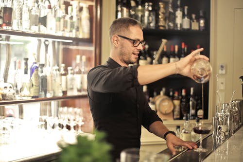 Free Side view of positive young barman in black uniform and eyeglasses pouring wine into glass while working at bar counter in modern restaurant Stock Photo
