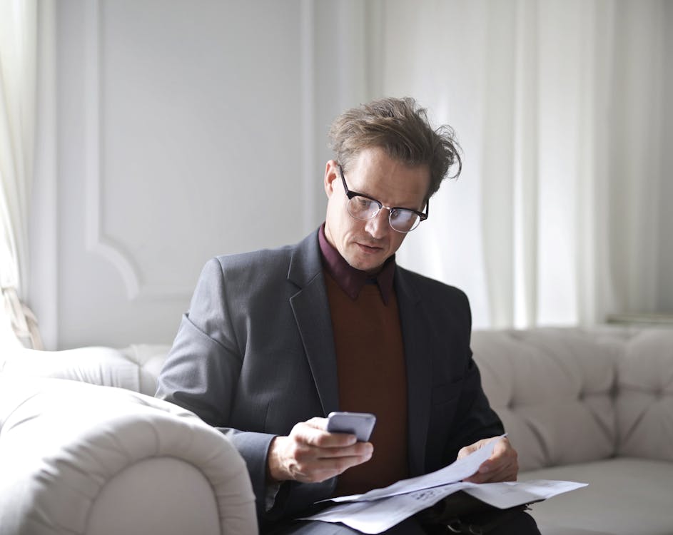 Free Photo of Man in a Suit and  Black Framed Eyeglasses Sitting on White Sofa While Using a Phone Stock Photo