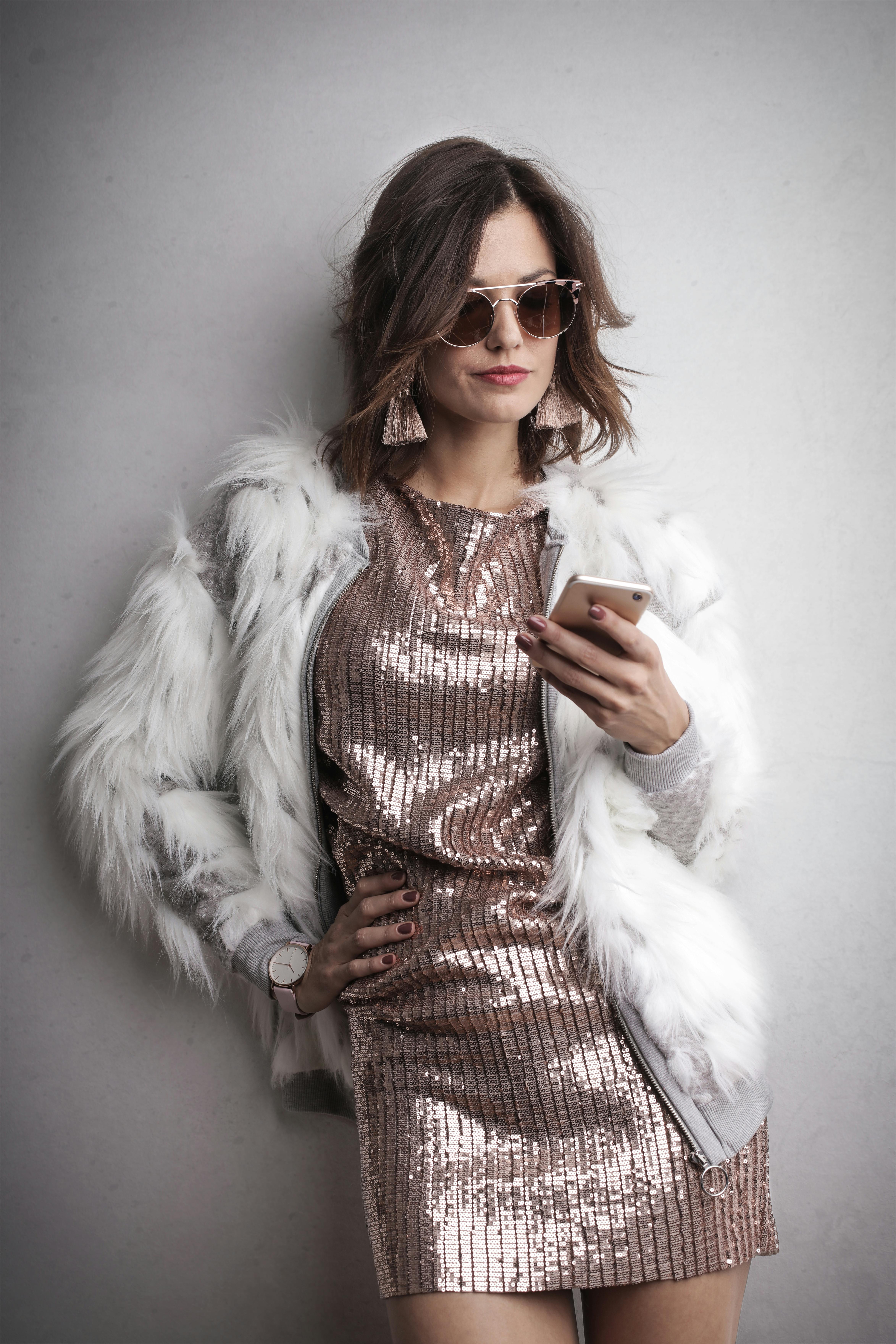 一位穿着亮片连衣裙和毛皮大衣的时尚女性，正靠在灰色墙壁上用智能手机发短信