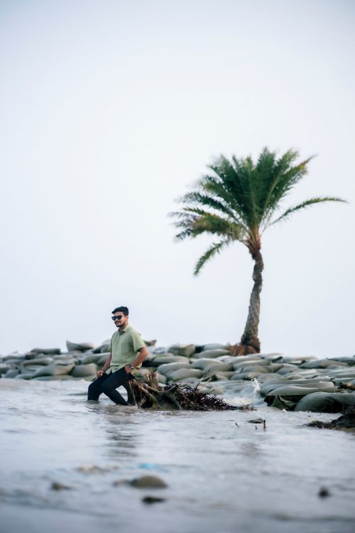 Free A man stands by a palm tree on a rocky coastline, casual and serene. Stock Photo