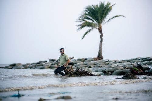 Free A man stands at the beach shore with a palm tree and sandbags in the background. Stock Photo